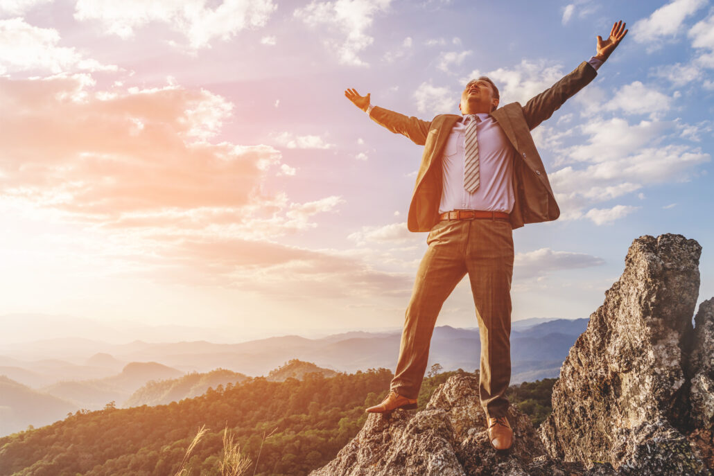 Businessman in a suit standing triumphantly on a mountain peak with arms raised, symbolizing achievement and financial freedom under a bright sky.