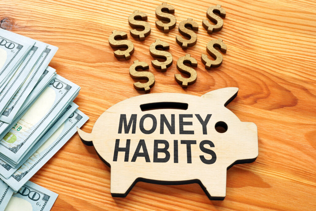 Wooden piggy bank labeled “Money Habits” next to stacks of hundred-dollar bills and wooden dollar signs on a wooden table surface, symbolizing financial habits and savings.