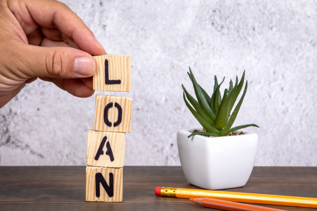 A hand stacking wooden blocks spelling the word "LOAN" next to a small potted plant and pencils, symbolizing financial decisions and rebuilding.