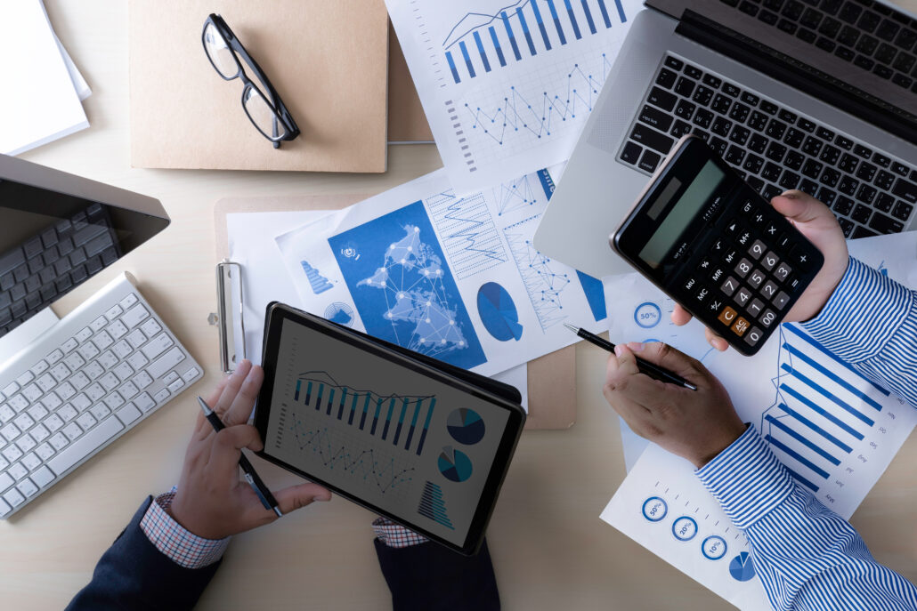 Overhead view of two people reviewing charts with a tablet, calculator, and laptop on a desk.