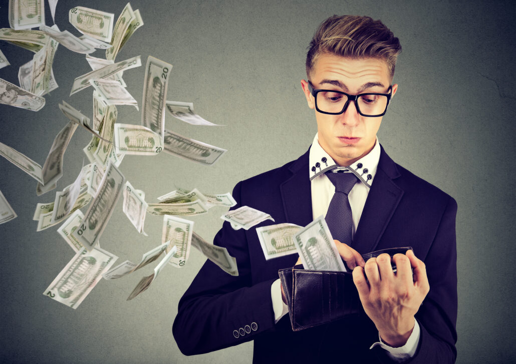 Young professional in a suit looks at his wallet while U.S. dollar bills scatter in the air.