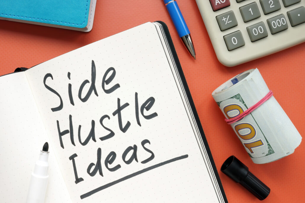 Overhead view of a notebook reading “Side Hustle Ideas” next to a calculator, pen, and a roll of cash.