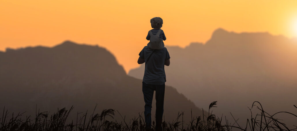 Silhouette of a parent carrying a child on their shoulders at sunset with mountains in the background.