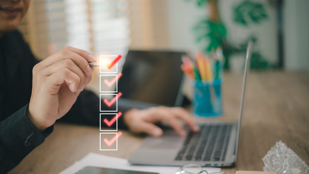 Person marking off items on a checklist while working on a laptop, symbolizing a simple monthly saving and investing routine.