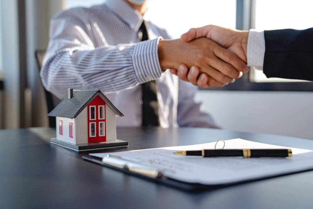 Two professionals shaking hands across a desk with a small model building and contract documents, representing a medical practice leasing property from a physician-owned real estate LLC.